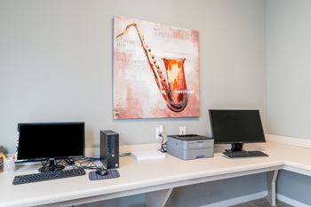A white desk with a computer, monitor, and a painting of a saxophone on the wall.
